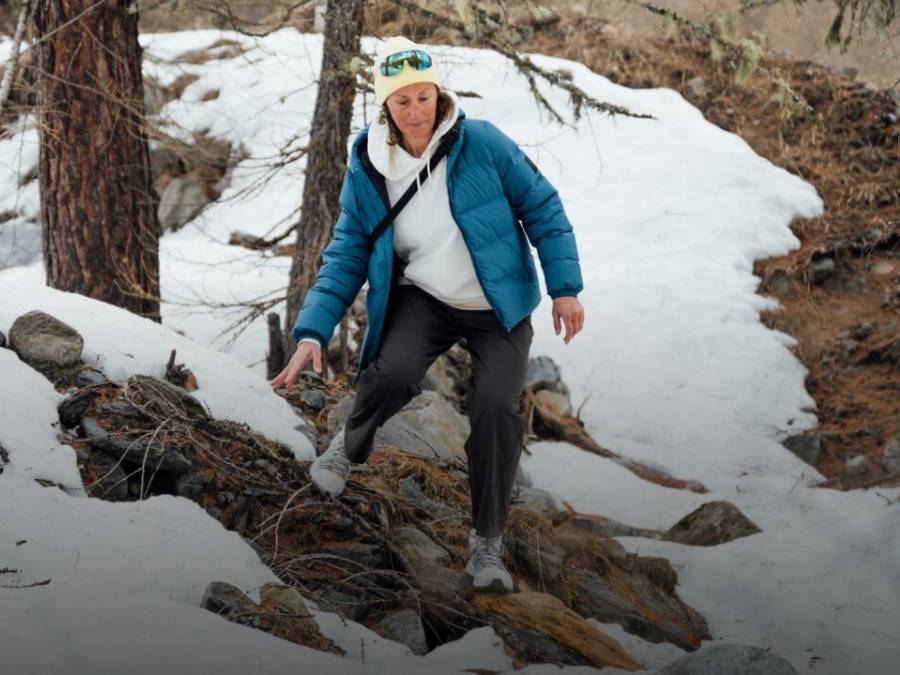 Twee sleeën met elk twee personen glijden een besneeuwde helling af. De mensen lachen en dragen winterkleding, mutsen en zonnebrillen; op de achtergrond zijn met sneeuw bedekte bergen te zien.