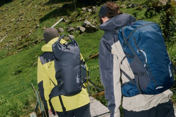 Twee wandelaars van achteren met rugzakken op hun rug die over een houten brug lopen