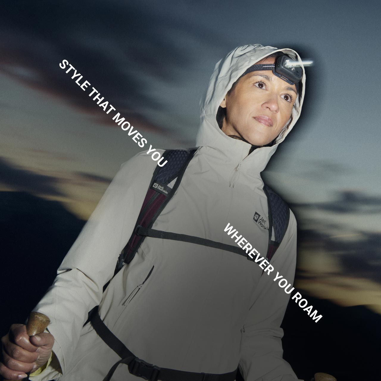 Iemand in een outdoor-uitrusting met een hoofdlampje kijkt vooruit en houdt trekkingstokken vast terwijl de avond valt in de bergen. De tekst luidt: "Style that moves you wherever you roam"