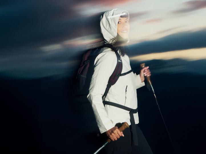 Iemand in wandelkleding, inclusief wit jack en capuchon, glimlacht terwijl ze trekkingstokken vasthoudt. De nevelige avondschemering suggereert beweging en avontuur.