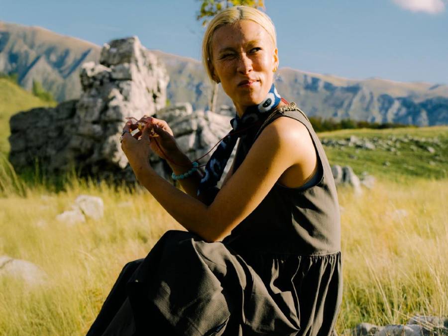 Een blonde vrouw zit in een groene weide en kijkt in de verte, omgeven door bergen