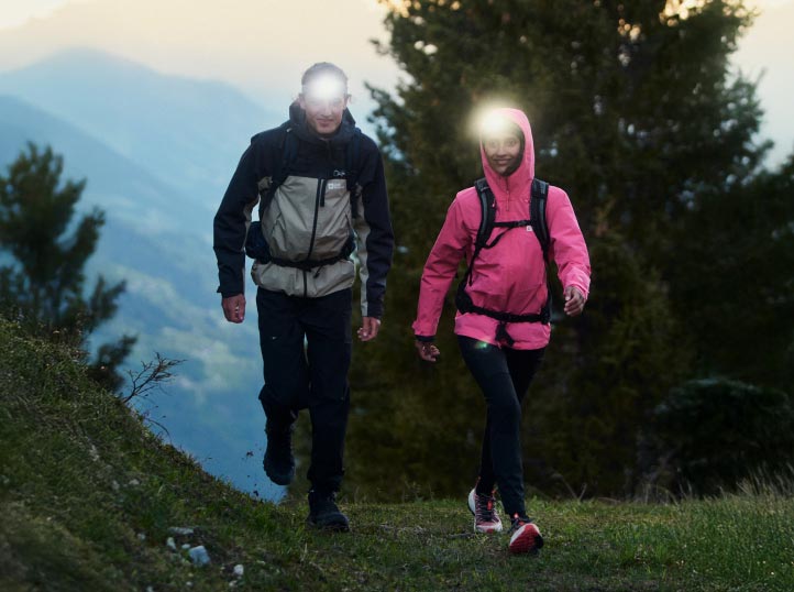 Twee wandelaars met hoofdlampjes lopen in de schemering op een bospad. Ze dragen jacks, de een zwart en grijs, de ander roze.
